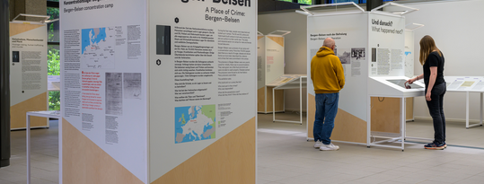 Ein Blick auf Ausstellungselemente der Sonderausstellung "Ein Tatort: Bergen-Belsen". Zwei Personen besichtigen die Ausstellung. Der Mann liest gerade einen Text auf einem Ausstellungselement. Die Frau benutzt eine Medienstation.
